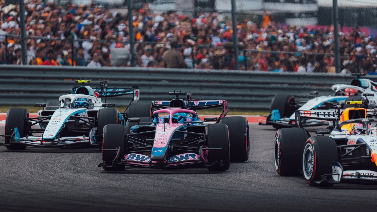 Monopost Alpine F1 al lui Pierre Gasly pe pistă în Marele Premiu al Statelor Unite 2026, surprins din față în timpul unei curbe, urmat de alte monoposturi și cu tribune pline în fundal. Culoare albastru-roșu, unghi joasă apropiat de asfalt.