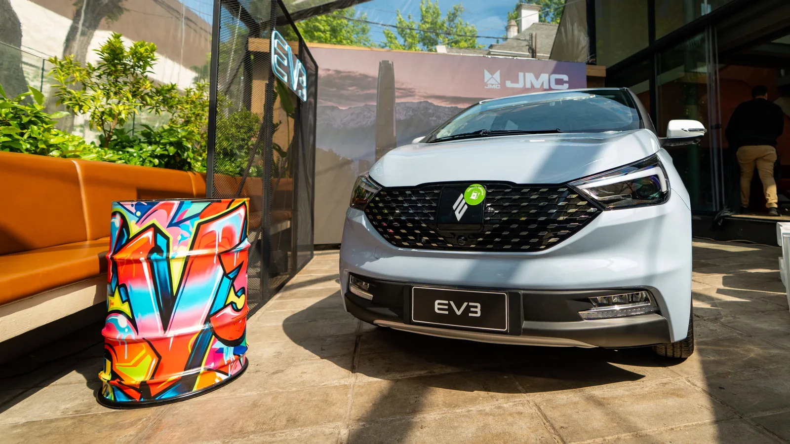 Partea frontală a unui JMEV EV3 alb, fotografiat în exterior, în fața unui spațiu cu geamuri mari și bannere cu logo-ul JMEV. Mașina are lumini LED și grilă neagră cu elemente distinctive. În dreapta, o valiză colorată cu model geometric stă pe trotuar.