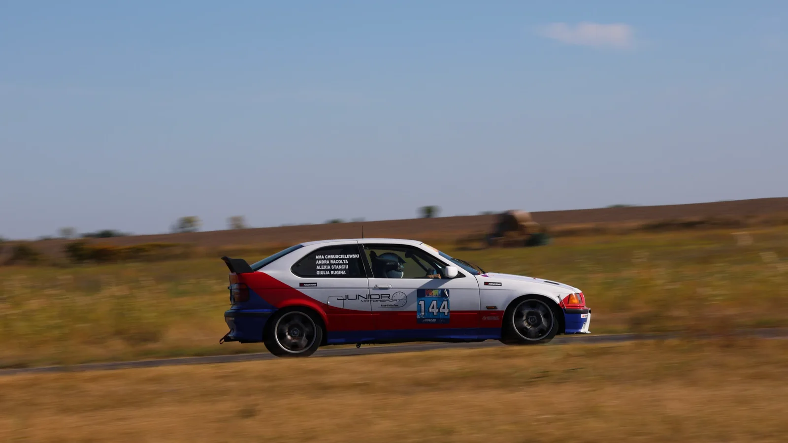BMW Seria 3 E36 de curse, cu numărul 144, rulează pe un traseu de rally-raid printr-un peisaj deschis.
