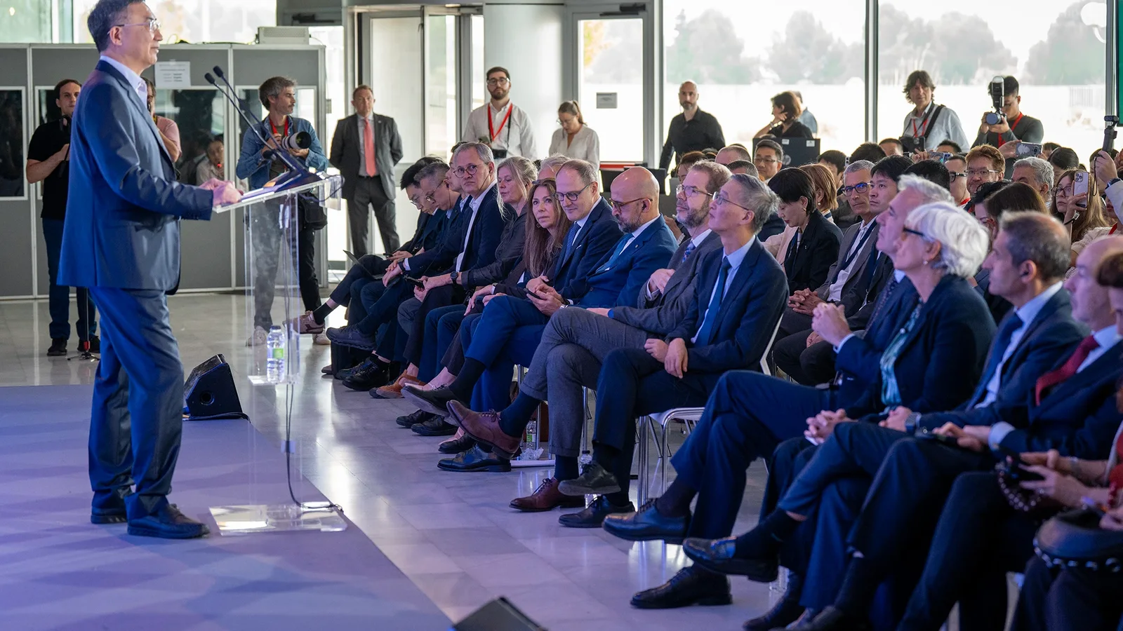 Bărbat în costum transmite un discurs în fața unui audiențe formate din persoane în ținute business, așezate pe scaune într-o încăpere luminoasă cu ferestre mari, eveniment oficial de prezentare sau conferință, perspectivă din lateral și ușor de jos.