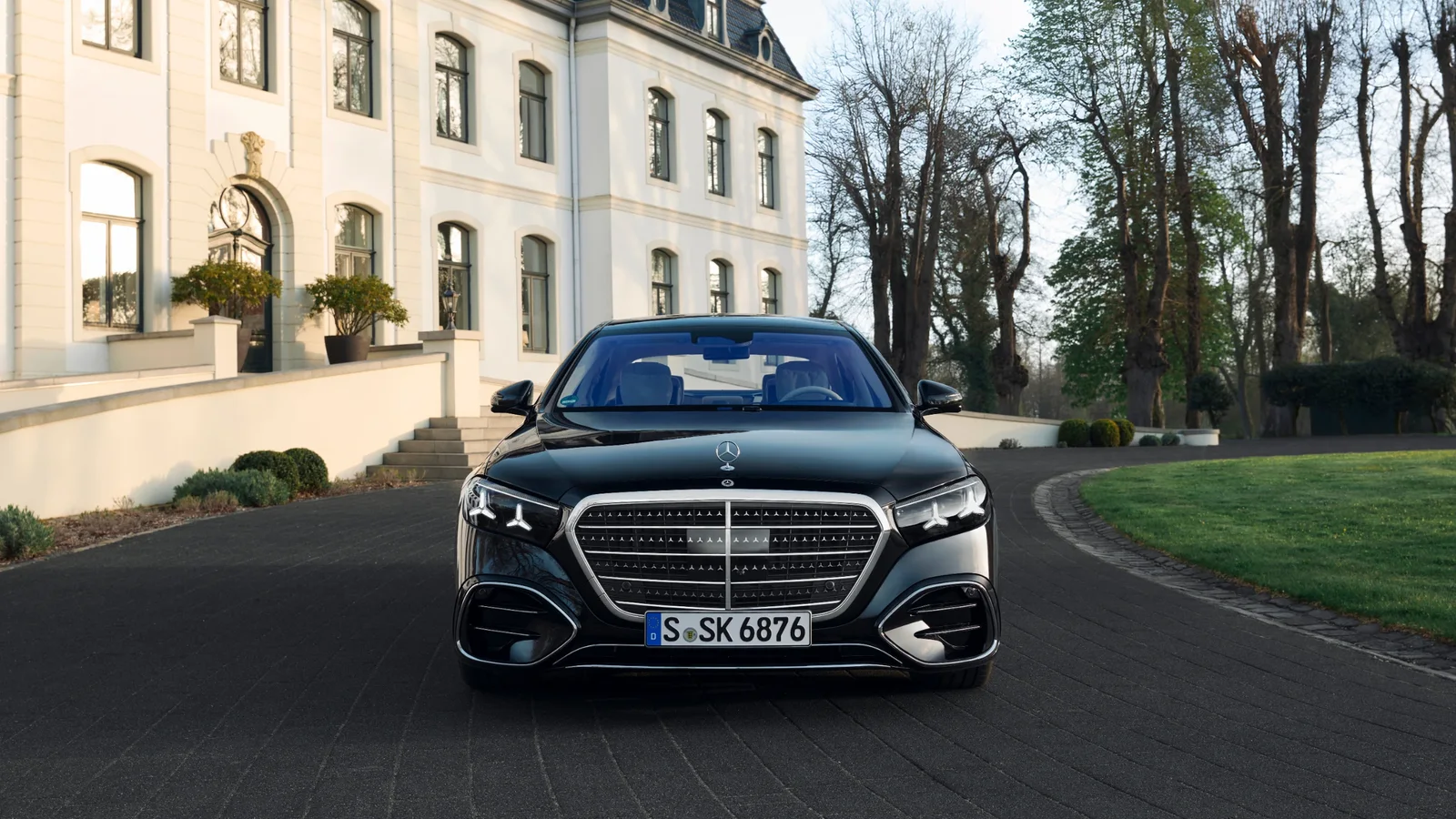 Mercedes-Benz Clasa S neagră văzută frontal pe o alee în fața unei clădiri elegante albe cu ferestre înalte, iluminată natural, într-un cadru urban cu copaci și spații verzi; fotografia evidențiază designul exterior sofisticat și grila frontală distinctivă.