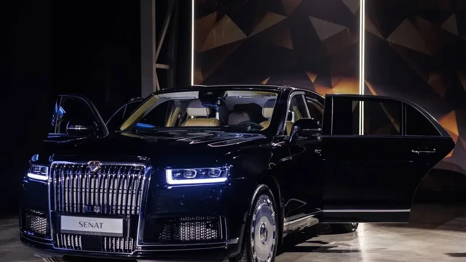 Aurus Senat modernizat, sedan negru cu faruri Matrix LED aprinse în pavilion interior, ușile din față și spate deschise, vizibilă grila frontală mare și lucioasă, imagine realizată din față și lateral dreapta, fundal întunecat cu elemente geometrice.