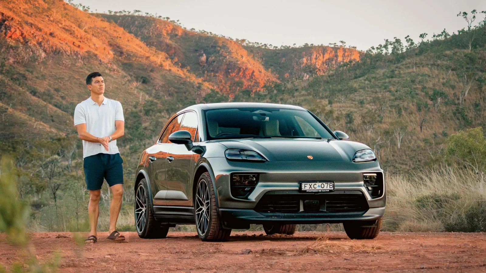 Porsche Macan 4S gri închis parcat pe un drum de pământ roșu, cu un bărbat în picioare lângă mașină. Fundal cu dealuri joase și vegetație specifică deșertică, lumină caldă de apus sau răsărit, fotografie realizată din față, ușor lateral.