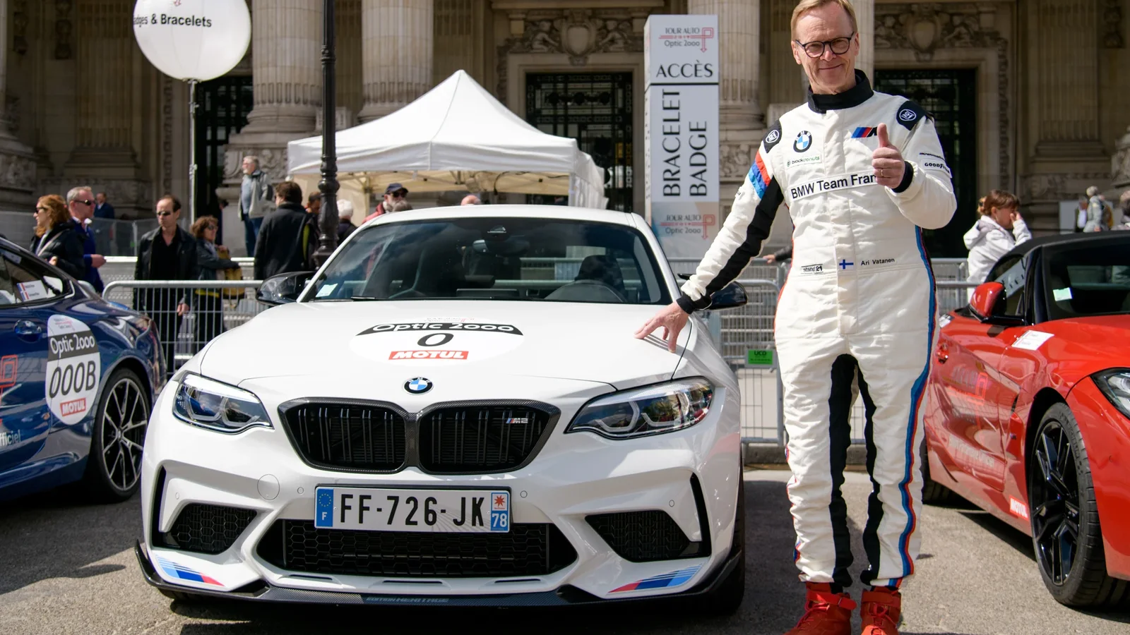 Ari Vatanen în combinezon alb de curse, stând lângă un BMW alb parcat frontal, sub o cortină albă, pe fundal de clădire monumentală și mulțime de oameni la Trofeul Poiana Brașov. Alături, se vede o mașină sport roșie.