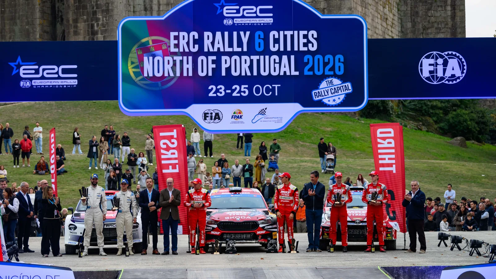 Startul unei curse de raliu ERC în Portugalia, cu o echipă de piloți și mașina Lancia Ypsilon Rally2 HF Integrale aliniate în linie, în fața unei panouri albastre care indică „ERC Rally 6 Cities North of Portugal 2026”. Cer senin, suprafață asfaltată, participanți în costume de curse.