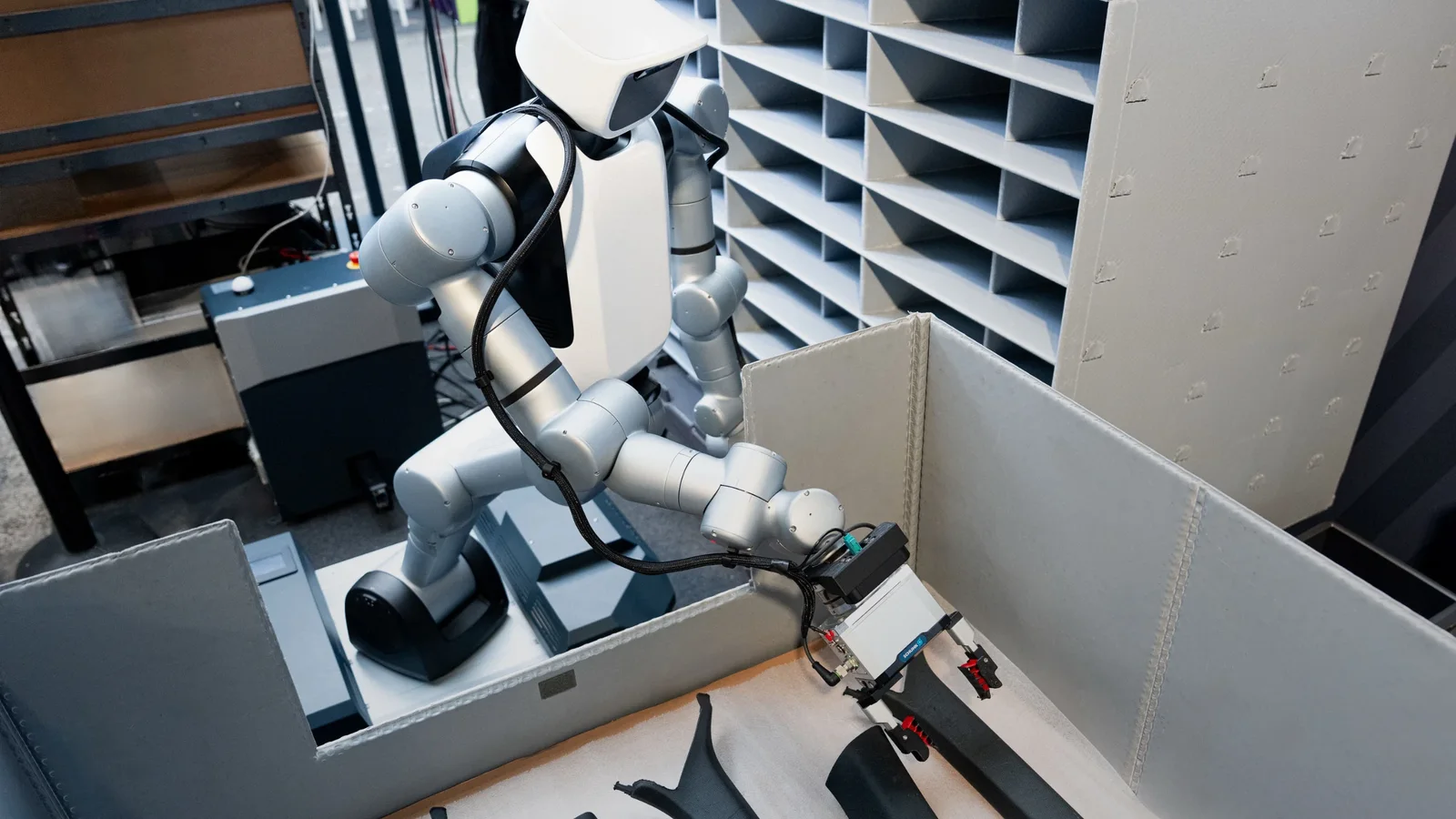 Robot umanoid mobil cu două brațe, de culoare albă și neagră, poziționat lângă un sistem de rafturi albe pentru piese, ridicând un obiect mic, fotografia este realizată din plan apropiat, într-un spațiu industrial luminat artificial.