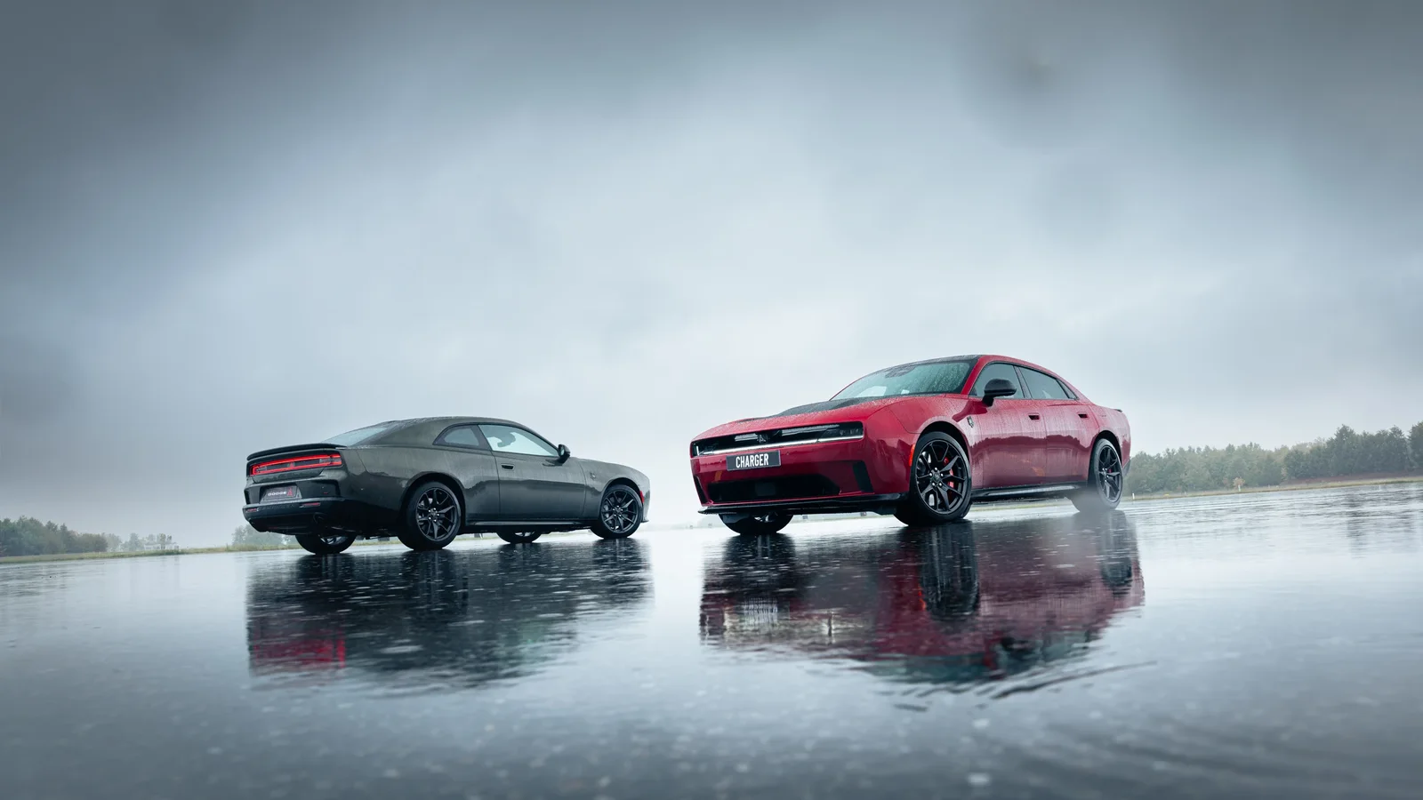 Două modele Dodge Charger, unul gri și unul roșu, sunt fotografiate pe o suprafață umedă care reflectă mașinile, sub un cer noros. Vedere laterală și frontală, pe un fundal natural neclar, într-un cadru larg, prezentând designul sportiv al mașinilor.