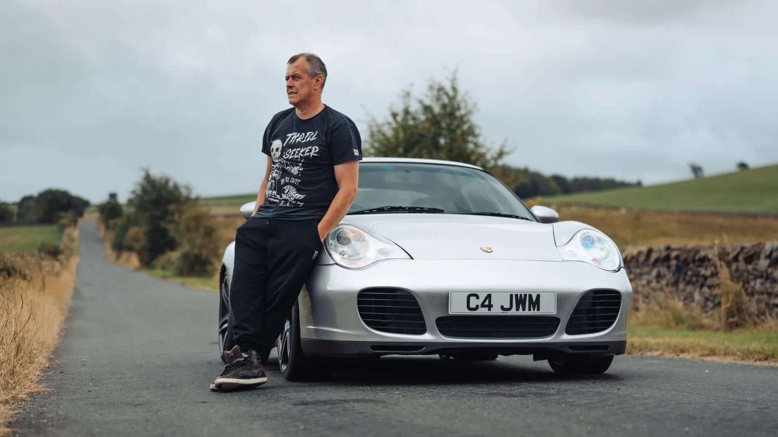 John McGuinness stă relaxat sprijinit de capota unui Porsche 911 Carrera 4S din generația 996, parcat pe un drum rural asfaltat. Fundalul include peisaj cu câmpuri verzi și un cer noros, fotografia este realizată din față și unghi jos, în culori naturale.