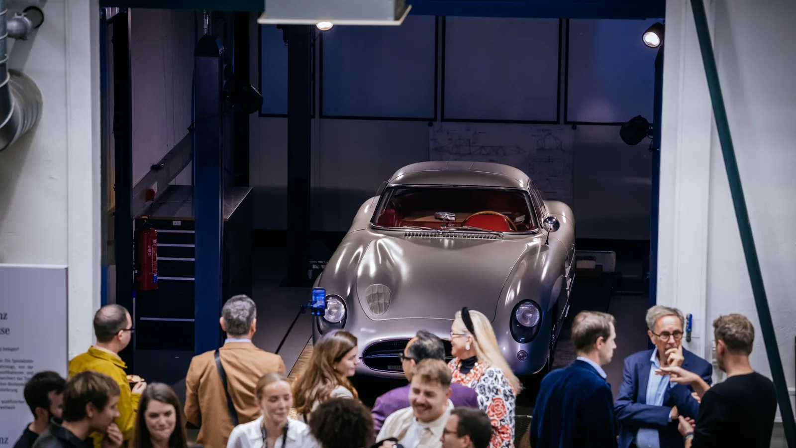 Mașină Mercedes-Benz clasică, argintie, expusă într-un spațiu interior al centrului Classic Center Fellbach, iluminare artificială albă, în față oameni adunați privind și discutând despre vehicul.