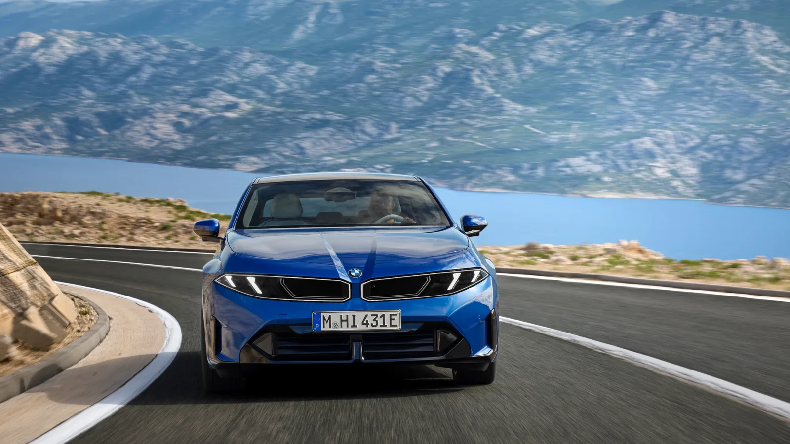 BMW albastru, văzut din față, pe un drum de coastă cu lac și munți în fundal.