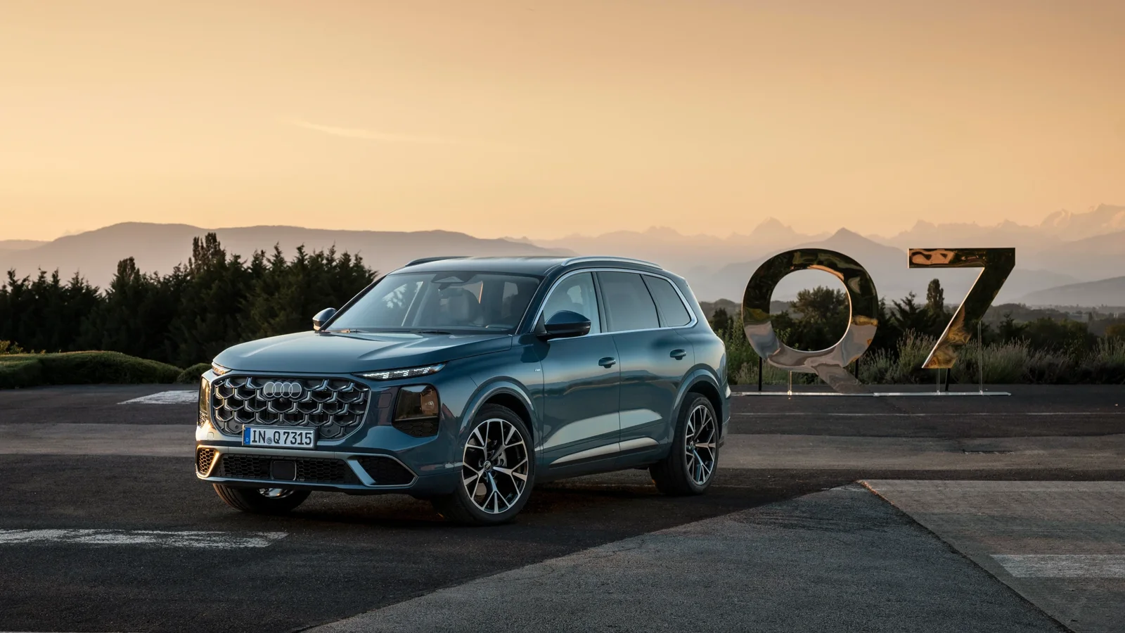 Audi Q7 albastru fotografiat pe un platou la apus, cu munți în fundal și o instalație „Q7” în partea dreaptă.