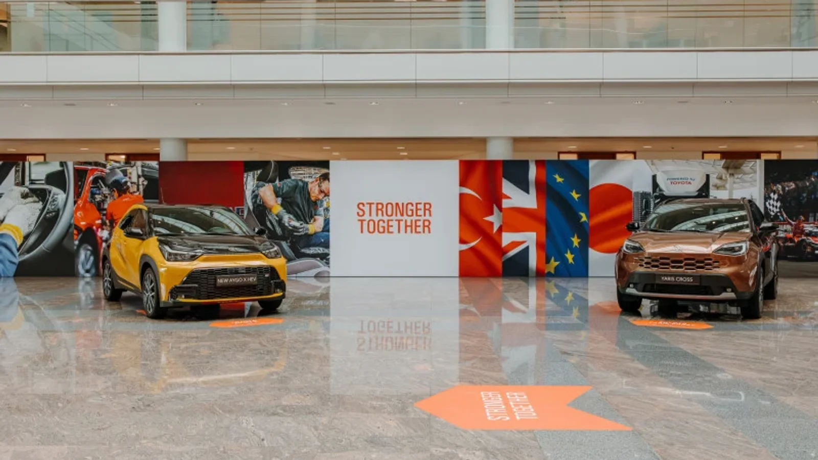 Interior spațios cu două Toyota Yaris, una galbenă și una maro, expuse față în față pe un podea lucioasă; în centru, un panou alb cu text roșu „Stronger Together”; fundal cu pereți colorati și geamuri mari, iluminare naturală.
