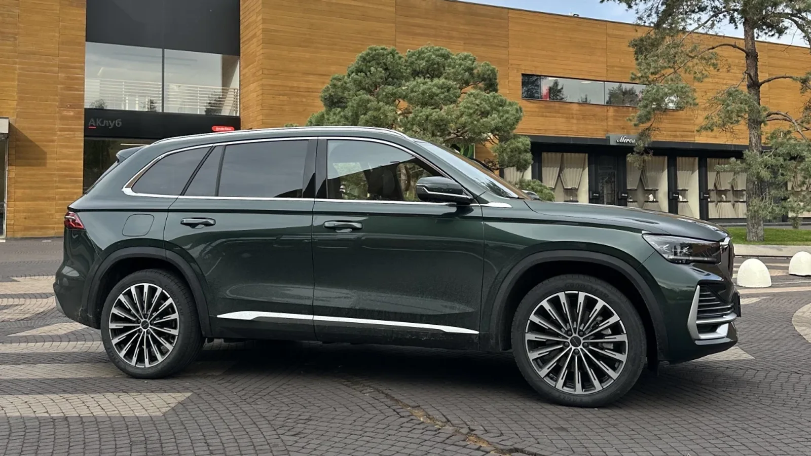 SUV Volga K50 de culoare verde închis, văzut lateral, oprit pe pavaj în fața unei clădiri moderne cu fațadă din lemn și geamuri largi, sub un cer senin și printre câțiva copaci.