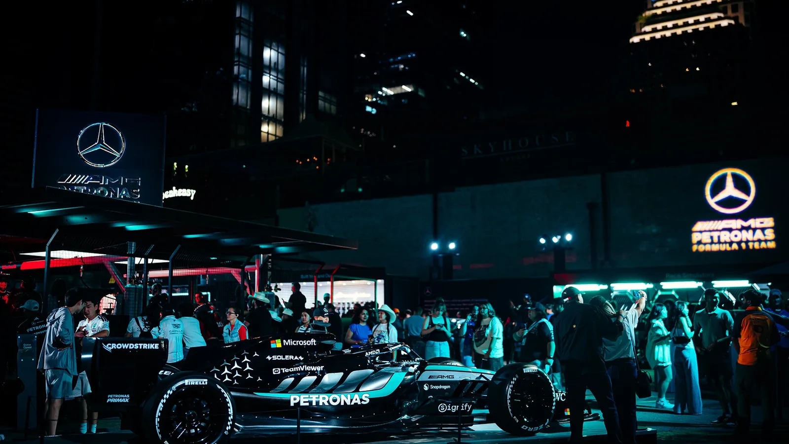 Monopost Mercedes-AMG PETRONAS F1 2026 expus într-un Fan-Hub Mercedes la Las Vegas noaptea, cu mulțime de oameni în jur, iluminare albastru-verzuie și clădiri în fundal. Logo-uri Mercedes și branding AMG vizibile pe ecrane și panouri.