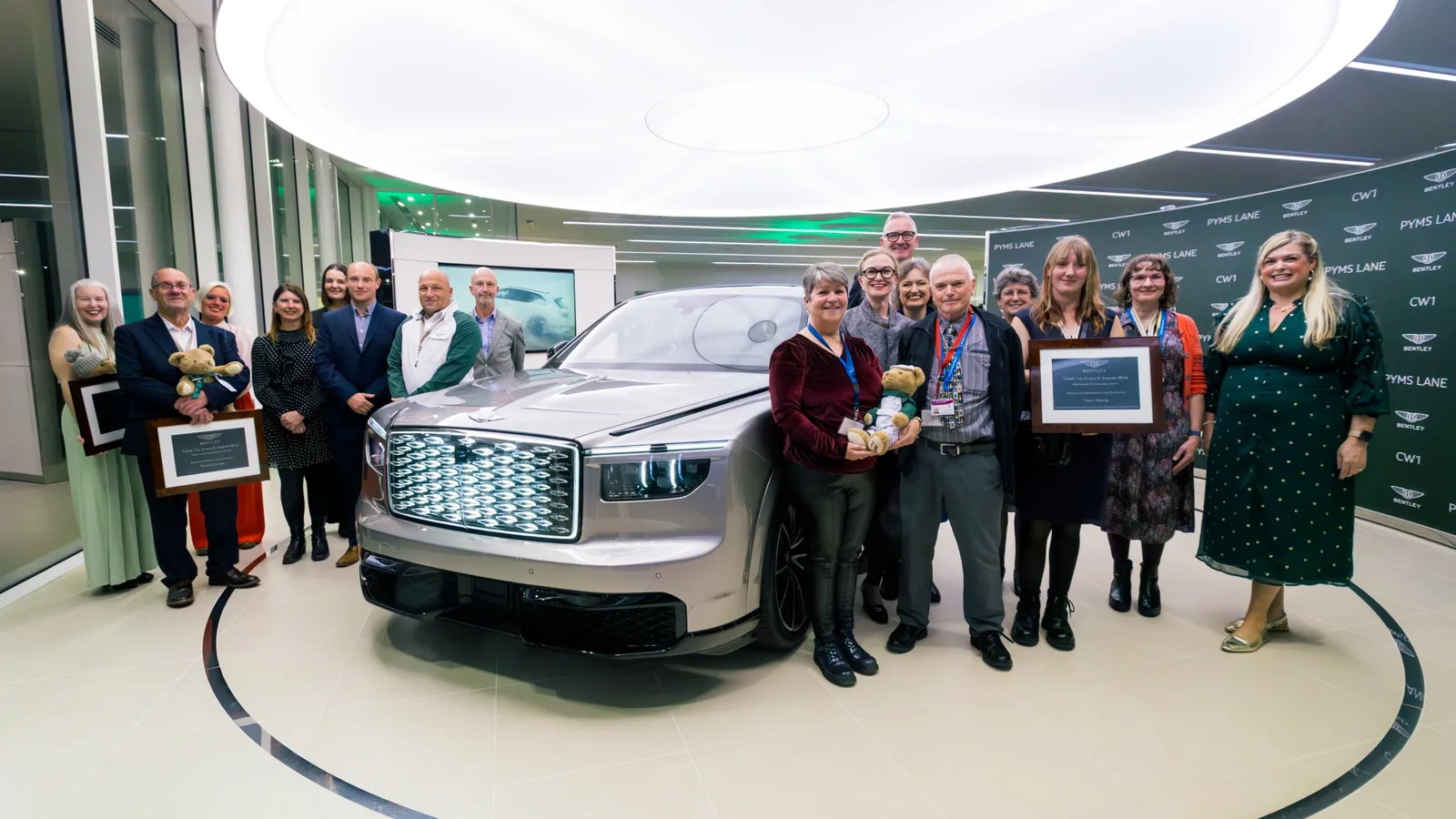 Grup de aproximativ 15 persoane, majoritatea zâmbitoare, pozând în jurul unui automobil Bentley în spațiu interior modern, cu podea și pereți albi, iluminat puternic de o lumină circulară de tavan. Unele persoane țin trofee și rame cu diplome.