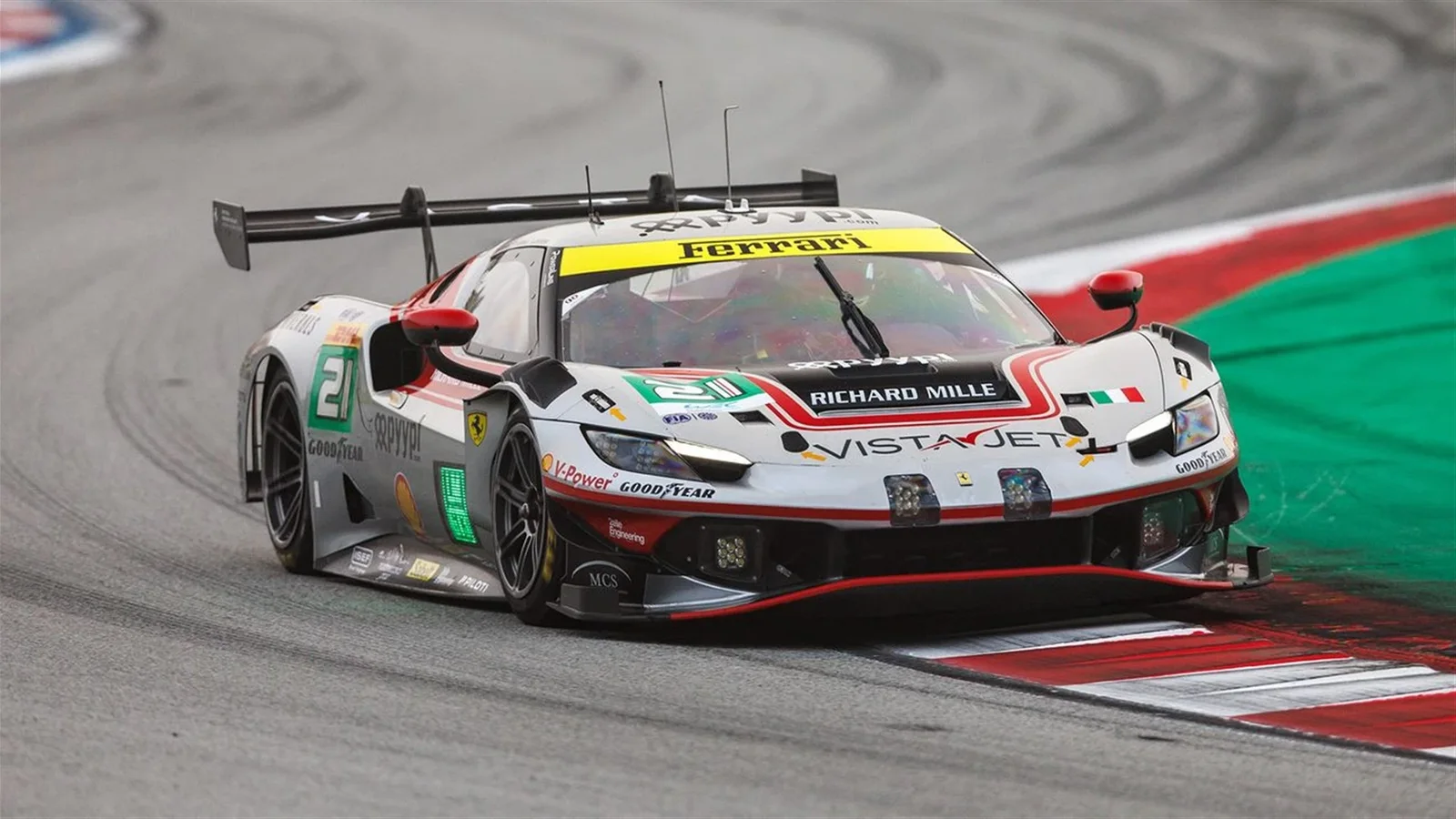 Ferrari 296 LMGT3 Evo numărul 21 în viraj pe circuitul de la Barcelona, văzut din față-oblic, cu culoare alb-roșu-verde și elemente grafice distinctive, pe linia de bordură roșie și albă a pistei în sesiunea de calificări FIA WEC.