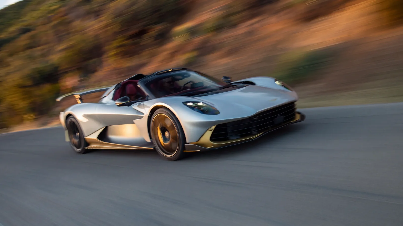 Aston Martin Valhalla Spider argintiu, decapotabil, în viraj pe un drum asfaltat, fotografie realizată din lateral-spate, fundal natural defocalizat, mașina în mișcare.