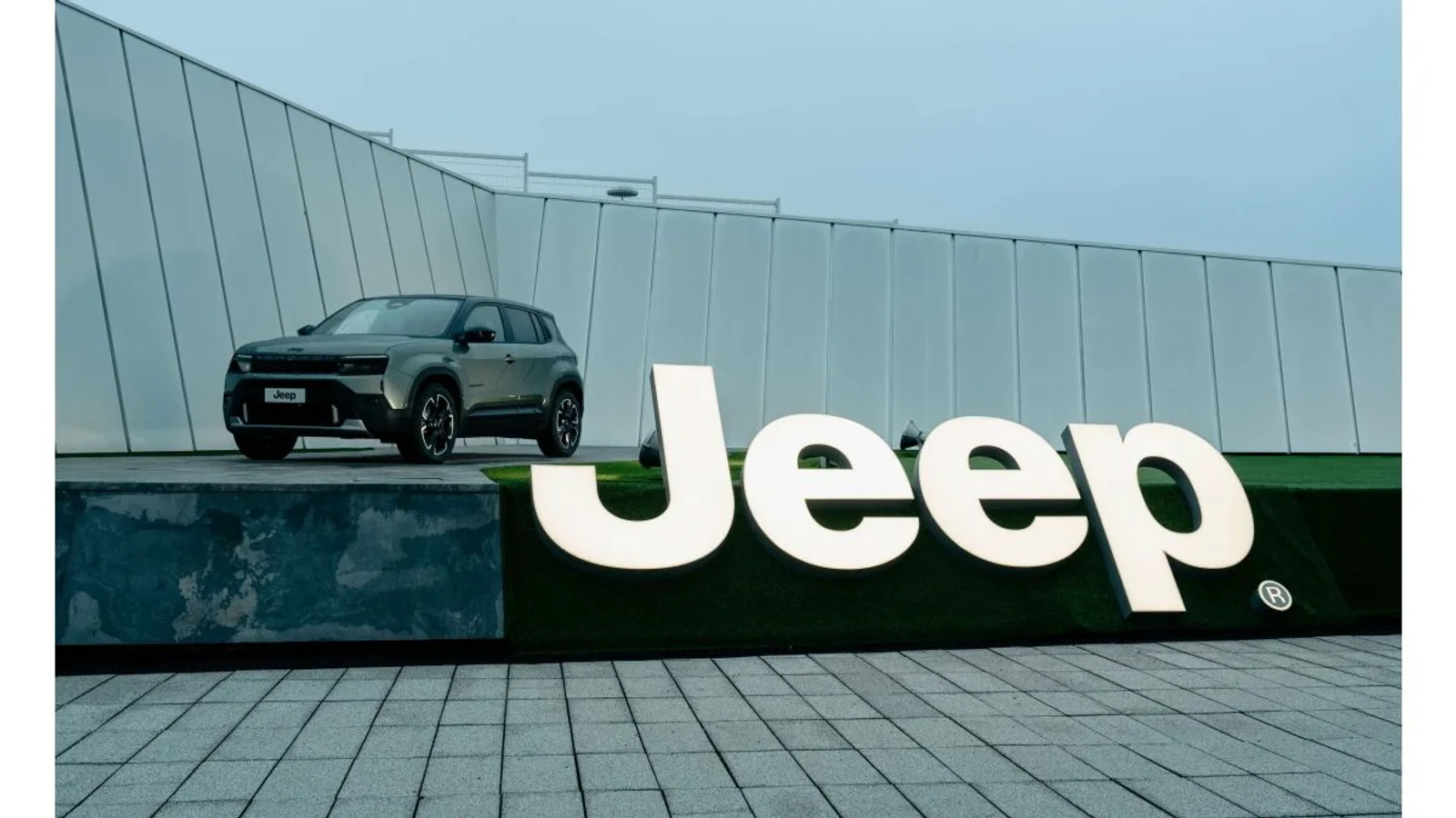 Mașina Jeep Avenger de culoare închisă este expusă pe acoperișul unei clădiri moderne, transparentă, cu panouri de sticlă, în spatele unui panou mare cu litere albe ce formează logo-ul Jeep, pe fundal cer senin și lumină naturală difuză.