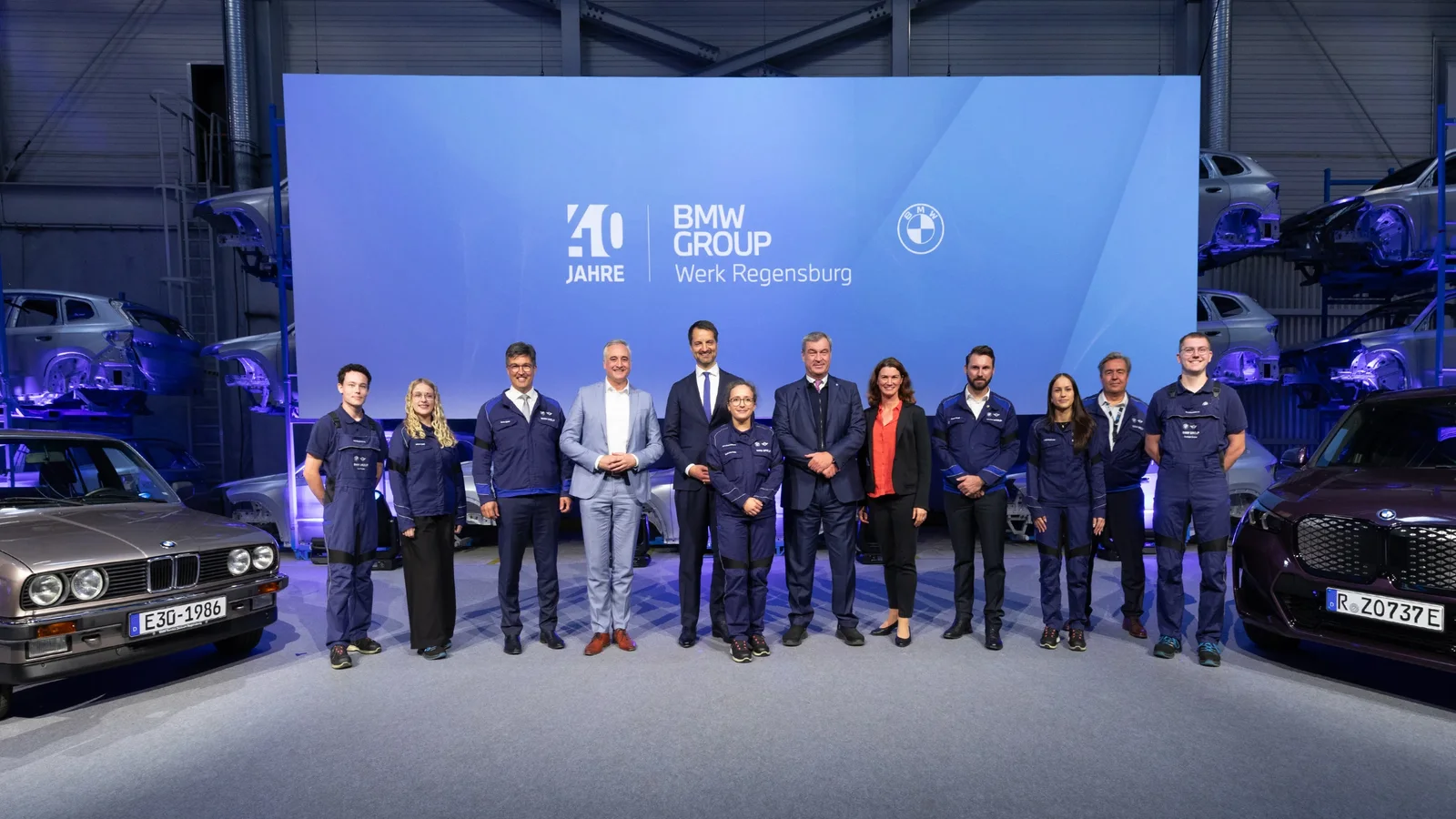 Grup de unsprezece persoane, majoritar bărbați în ținută oficială sau casual, pe scenă la eveniment BMW Group din Regensburg, cu două BMW-uri clasice în lateral, pe fundal albastru cu logo-urile BMW și text aniversar pentru 40 de ani și nouă milioane de automobile produse.