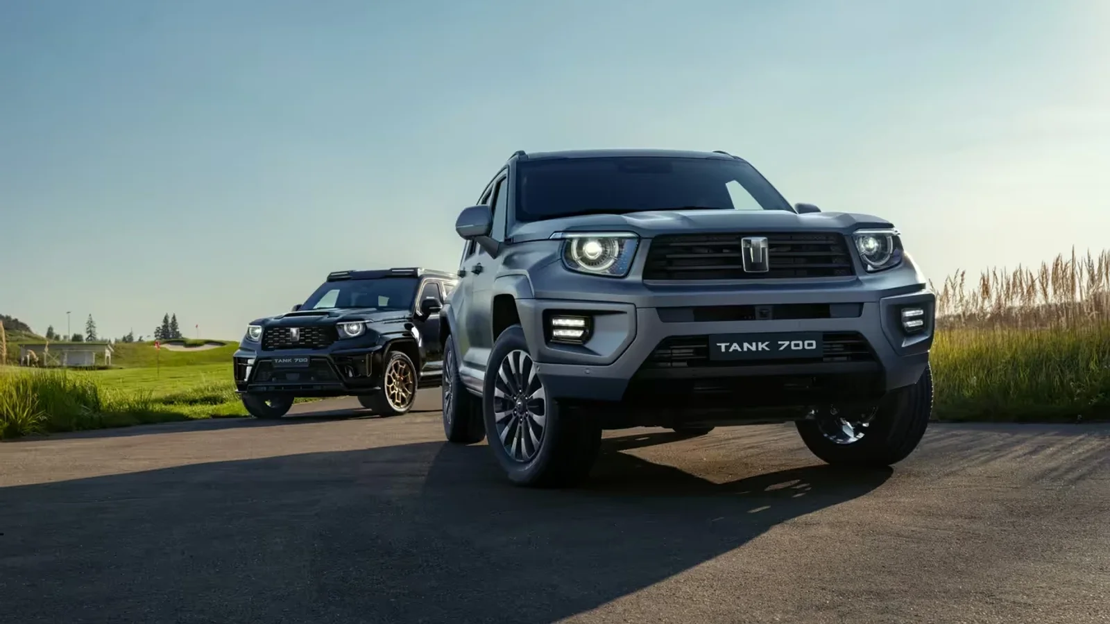 Două SUV-uri TANK de culoare gri și negru pe un drum asfaltat în lumina zilei, cu un fundal de iarbă și cer senin, fotografiate dintr-un unghi frontal-jos spre dreapta, evidențiind designul robust și farurile aprinse.