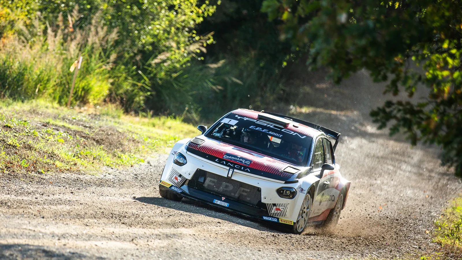 Lancia Ypsilon Rally2 a lui Nikolay Gryazin pe un drum de macadam, ridicând praf într-o zonă împădurită.